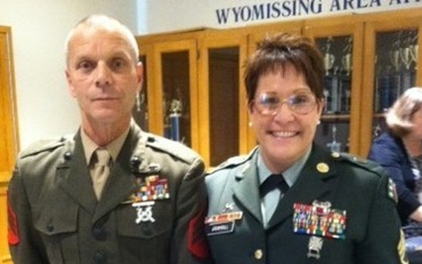 Two military personnel in dress uniforms pose together indoors at what appears to be a formal military or school event. The individual on the left wears a United States Marine Corps dress uniform, while the individual on the right wears a United States Army dress green uniform. They are standing in front of a display case with a banner reading 'Wyomissing Area' visible in the background.