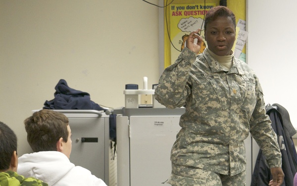 A U.S. Army soldier in ACU (Army Combat Uniform) is presenting or speaking to a group of students in a classroom setting. The soldier appears to be engaged in a community outreach or educational visit, addressing seated students at individual desks. A motivational poster reading 'If you don't know, ASK QUESTIONS' is visible on the wall behind the soldier.