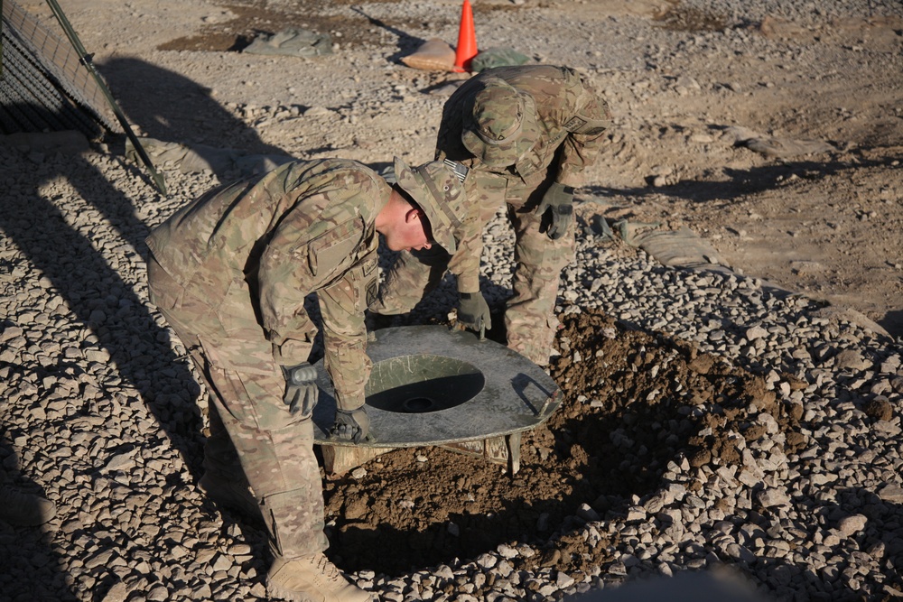 Two military personnel in MultiCam/OCP camouflage uniforms are installing or positioning what appears to be a field latrine seat/toilet over an excavated pit in a gravel-covered area. The soldiers are wearing tactical gloves and are bent over the hole, carefully placing the toilet seat fixture. An orange safety cone and HESCO barriers are visible in the background, suggesting a forward operating base or military installation.