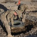 Two military personnel in MultiCam/OCP camouflage uniforms are installing or positioning what appears to be a field latrine seat/toilet over an excavated pit in a gravel-covered area. The soldiers are wearing tactical gloves and are bent over the hole, carefully placing the toilet seat fixture. An orange safety cone and HESCO barriers are visible in the background, suggesting a forward operating base or military installation.