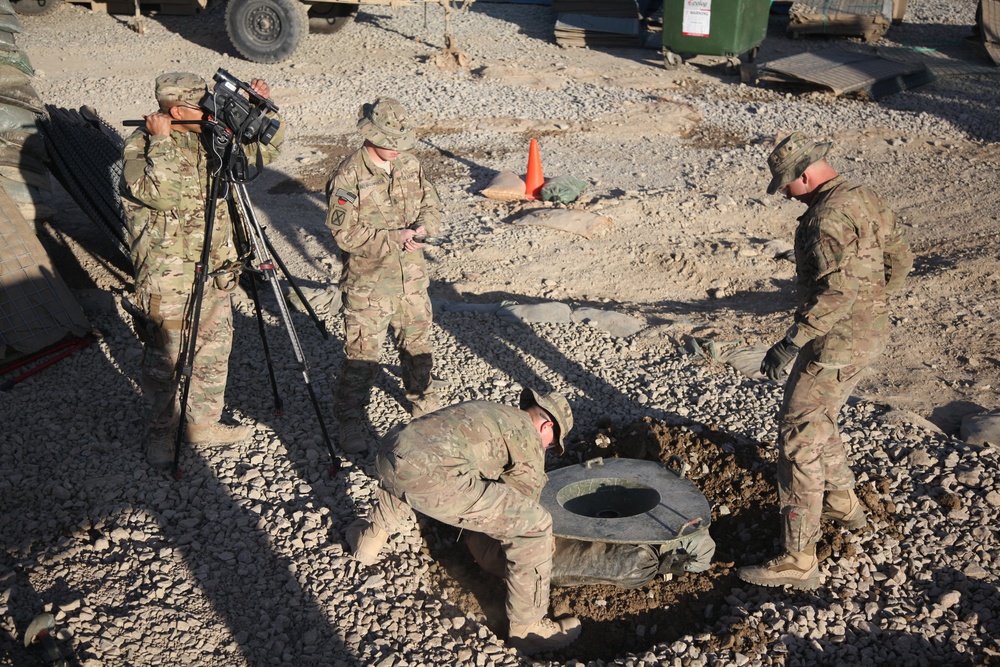 Four military personnel in MultiCam/OCP camouflage uniforms are engaged in what appears to be a maintenance or inspection activity at a forward operating base or military installation. One soldier is operating a video camera on a tripod, documenting the work, while another soldier is bent down inspecting or working inside a ground-level access point or utility hole. The setting is a gravel-covered outdoor area with military vehicles and equipment visible in the background.