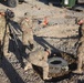 Four military personnel in MultiCam/OCP camouflage uniforms are engaged in what appears to be a maintenance or inspection activity at a forward operating base or military installation. One soldier is operating a video camera on a tripod, documenting the work, while another soldier is bent down inspecting or working inside a ground-level access point or utility hole. The setting is a gravel-covered outdoor area with military vehicles and equipment visible in the background.