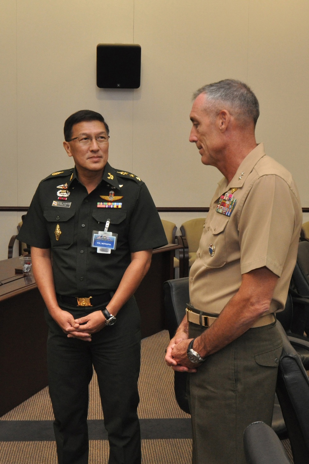 Two military officers in uniform are engaged in conversation in what appears to be a conference room setting. The officer on the left wears a dark green uniform with a visible name badge reading 'COL. MOTHAPOL' and multiple decorations, consistent with a Thai military uniform. The officer on the right wears a tan/khaki U.S. Marine Corps uniform with rank insignia and service ribbons.