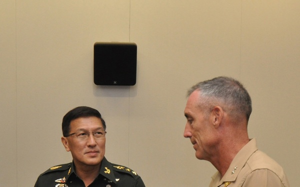 Two military officers in uniform are engaged in conversation in what appears to be a conference room setting. The officer on the left wears a dark green uniform with a visible name badge reading 'COL. MOTHAPOL' and multiple decorations, consistent with a Thai military uniform. The officer on the right wears a tan/khaki U.S. Marine Corps uniform with rank insignia and service ribbons.