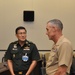 Two military officers in uniform are engaged in conversation in what appears to be a conference room setting. The officer on the left wears a dark green uniform with a visible name badge reading 'COL. MOTHAPOL' and multiple decorations, consistent with a Thai military uniform. The officer on the right wears a tan/khaki U.S. Marine Corps uniform with rank insignia and service ribbons.