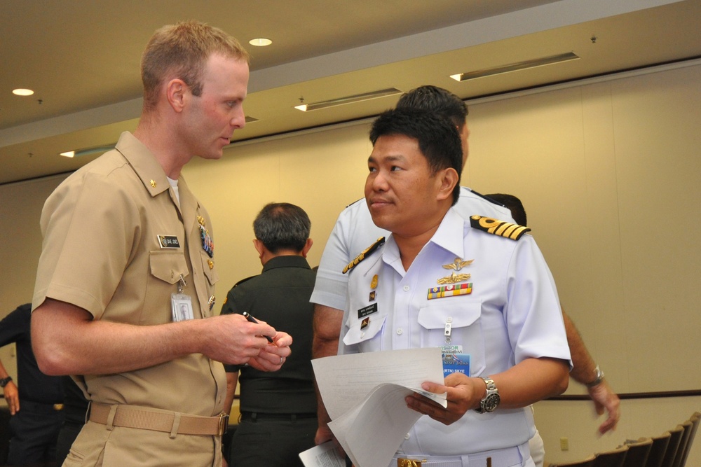 Two military officers in uniform are engaged in conversation at what appears to be a formal military conference or meeting. One officer is wearing a US Navy khaki service uniform, while the other is wearing a white naval uniform with gold shoulder boards and service ribbons, likely from a Southeast Asian navy. The setting appears to be an indoor conference facility.