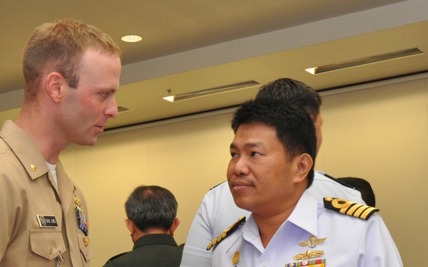 Two military officers in uniform are engaged in conversation at what appears to be a formal military conference or meeting. One officer is wearing a US Navy khaki service uniform, while the other is wearing a white naval uniform with gold shoulder boards and service ribbons, likely from a Southeast Asian navy. The setting appears to be an indoor conference facility.