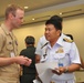Two military officers in uniform are engaged in conversation at what appears to be a formal military conference or meeting. One officer is wearing a US Navy khaki service uniform, while the other is wearing a white naval uniform with gold shoulder boards and service ribbons, likely from a Southeast Asian navy. The setting appears to be an indoor conference facility.