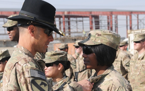 A U.S. Army officer wearing a black cavalry Stetson hat is pinning an award or insignia onto a female soldier during what appears to be a military ceremony. Both individuals wear MultiCam (OCP) uniforms with 1st Cavalry Division patches and ISAF (International Security Assistance Force) patches, indicating deployment to Afghanistan. A formation of soldiers stands in the background on a gravel surface near military structures.