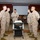 A group of U.S. Marines in desert MARPAT camouflage uniforms are gathered around a service cart with a white-frosted cake, likely during a military ceremony or celebration. A central figure appears to be holding a plate with a slice of cake, while others stand at attention or in relaxed postures. The setting appears to be an indoor dining or event facility with red walls, a projection screen, and an American flag visible in the background.