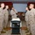 A group of U.S. Marines in desert MARPAT camouflage uniforms are gathered around a white cake on a rolling cart, appearing to participate in a military cake-cutting ceremony. A central figure is holding what appears to be a ceremonial sword or saber and is in the process of cutting the cake, which displays a red emblem consistent with the Marine Corps insignia. Additional personnel are seated at a table in the background, suggesting a formal military event or celebration.