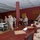 A military ceremony or formal gathering is taking place in an indoor venue with dark red walls and ornate curtains. Personnel in U.S. Marine Corps desert camouflage uniforms (MARPAT) are standing in formation on the left, while individuals in tan/khaki uniforms are seated at a long wooden table on the right, suggesting a review board or award ceremony setting. A service cart with what appears to be a cake is visible near the standing personnel.
