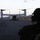 A military tiltrotor aircraft, identifiable as a Bell Boeing V-22 Osprey, is parked on a runway with its rotors spinning and rear cargo ramp lowered. A silhouetted military personnel member wearing a helmet is visible in the foreground right, observing the aircraft. The scene appears to take place at dusk or dawn at a remote or austere airstrip surrounded by sparse vegetation.