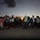 A large crowd of people, including adults and children, are gathered on an open paved area at dusk or dawn. They appear to be waiting, possibly at a port, airstrip, or evacuation point, with luggage and bags visible on the ground. The low light conditions and the presence of families with belongings suggest a mass movement or evacuation scenario.