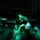 A dimly lit scene bathed in green light showing multiple individuals in close proximity. A central male figure appears distressed or in pain, with his hand raised to his forehead. The scene appears chaotic and crowded, possibly in an enclosed space.