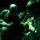 A bald adult male wearing a military-style jacket with a patch is seated and interacting with a young child in a darkened environment illuminated by green lighting. The child appears to be holding small yellow objects while the adult leans in attentively. The scene suggests a calm, personal interaction in what may be a military transport or low-light operational setting.