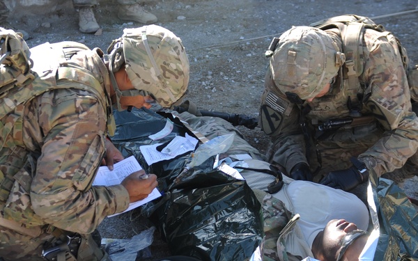 Two soldiers in MultiCam/OCP camouflage uniforms are providing tactical combat casualty care (TCCC) to a downed individual lying on a litter or tarp on the ground. One soldier appears to be documenting medical information on a card or form while the other attends to the casualty. Medical supplies and equipment are visible nearby, suggesting this is either a real casualty care scenario or a training exercise.
