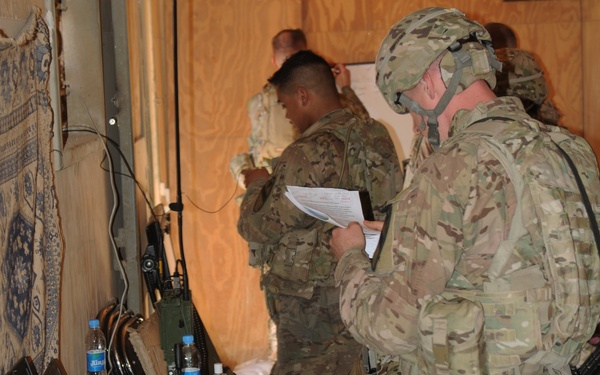 Military personnel in MultiCam/OCP camouflage uniforms are gathered in what appears to be a forward operating base or tactical operations center. Soldiers are reviewing documents and operating communications and computing equipment on folding tables. The setting suggests a briefing or mission planning session in a field environment.