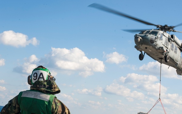 A military helicopter, appearing to be an MH-60 Seahawk, hovers over a naval vessel's flight deck while conducting a vertical replenishment (VERTREP) operation, lifting a cargo pallet via a sling load. A deck crew member wearing a green vest and helmet marked '9A' is directing the operation with outstretched arms. Rows of what appear to be ordnance or supply containers are lined up on the flight deck in the foreground.