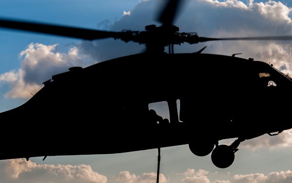 A military helicopter, identifiable by its silhouette as a UH-60 Black Hawk type, hovers low over a surface while two personnel in silhouette stand or move beneath it. A rope or cable extends from the helicopter toward one of the figures on the ground. The image is captured at dusk or dawn, creating a dramatic backlit silhouette composition against a partly cloudy sky.