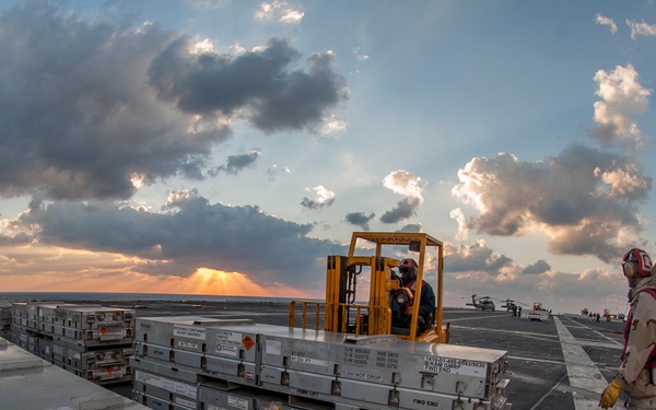 The image shows operations on the flight deck of a U.S. Navy aircraft carrier at sunset or sunrise. A forklift operator is maneuvering stacked military cargo containers while a crew member in a red helmet and flight deck gear observes and directs the operation. The wide-angle fisheye lens captures the expansive flight deck, superstructure, and dramatic cloud-filled sky over open ocean.