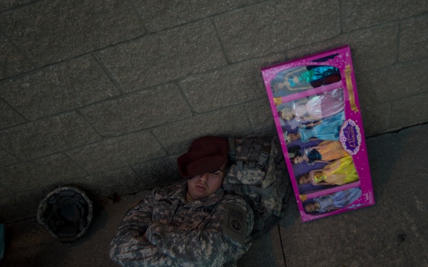 A soldier in camouflage uniform is lying on a concrete floor with arms crossed, appearing to be resting or sleeping. Nearby are children's toy boxes, including what appears to be a princess doll set in a pink box. The scene suggests a rest period during what may be a humanitarian aid or toy distribution mission.