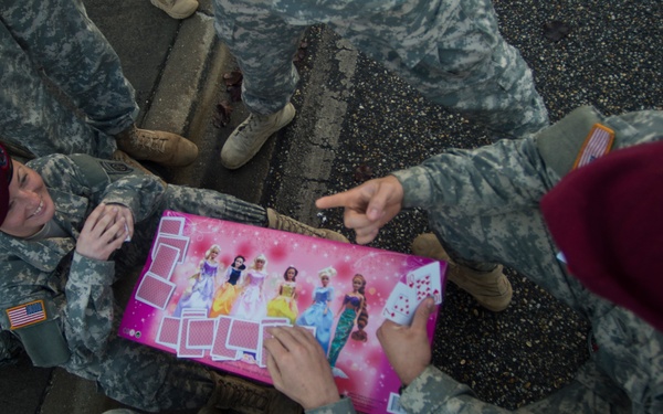 A group of soldiers in US Army camouflage uniforms and maroon berets are gathered on a paved surface, playing cards using a pink Disney Princess-themed card game mat or box. The soldiers appear to be relaxing during downtime, with one soldier smiling and others engaged with the cards. The maroon berets and '82nd Airborne' patch visible on at least one uniform suggest these are paratroopers.