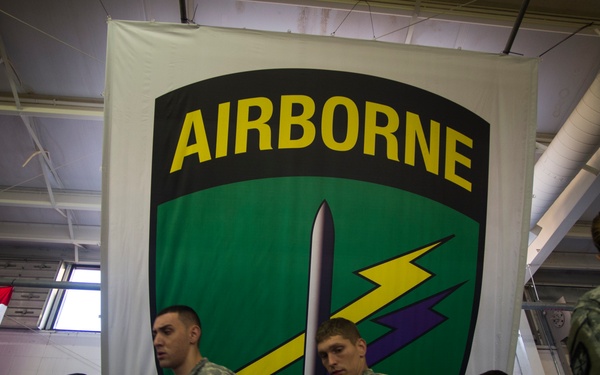 A group of U.S. Army soldiers in ACU (Army Combat Uniform) camouflage are gathered indoors beneath a large 'AIRBORNE' unit banner. The setting appears to be a charitable or community event, likely a toy drive, given the visible boxed toys and gifts on tables in the foreground. An American flag is partially visible in the background to the left.