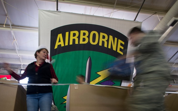 The image shows a military-related indoor event with an 'Airborne' unit banner prominently displayed in the background. Soldiers in camouflage uniforms are moving through what appears to be a distribution or processing area with large cardboard boxes. A civilian woman in casual clothing stands among the boxes, possibly assisting with the event.