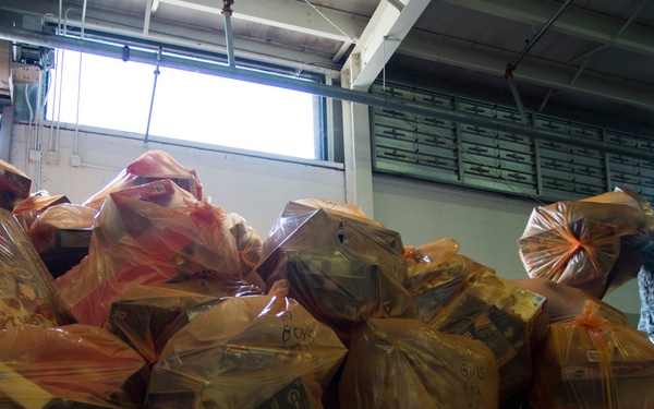A large pile of orange/yellow plastic bags filled with what appear to be toys and boxed items is stacked on a concrete floor inside a warehouse or gymnasium. A person in military camouflage uniform stands to the right, handling one of the bags. A bicycle is partially visible in the background, suggesting this may be a toy or gift collection/distribution event.