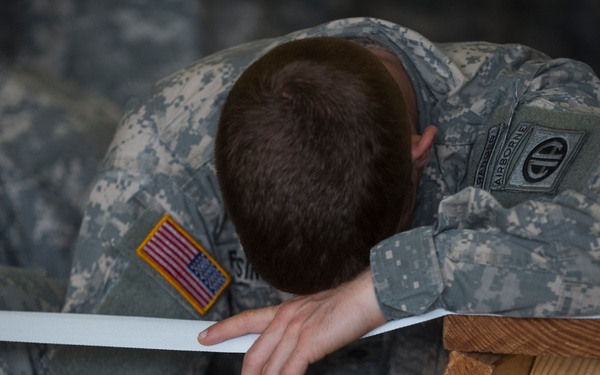 A U.S. Army soldier wearing ACU (Army Combat Uniform) with an 82nd Airborne Division patch is slumped forward with their head down on or near a wooden surface. The soldier appears to be resting or in a moment of exhaustion. A second soldier in similar uniform is partially visible in the background.