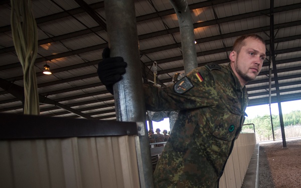 A soldier wearing German Bundeswehr camouflage uniform with a visible German flag patch is working inside a large covered structure, handling metal panels or barriers. The setting appears to be an industrial or agricultural facility with a corrugated metal roof. Other soldiers are visible in the background.