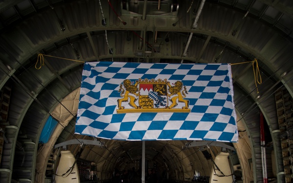 The interior of a large military transport aircraft, likely a C-130 Hercules, is shown with a Bavarian state flag (Freistaat Bayern) prominently displayed hanging from the fuselage ceiling. The flag features the distinctive blue and white diamond (lozenge) pattern with the Bavarian coat of arms in the center. A single person in a flight suit is visible in the background near the rear of the cargo hold.