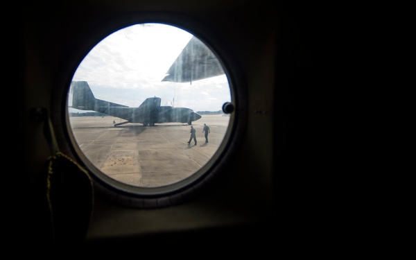 The image is taken from inside an aircraft looking through a circular porthole window onto a military flight line or tarmac. A large military transport aircraft, likely a C-130 Hercules, is visible parked on the concrete apron outside. Two personnel are walking on the tarmac near the aircraft, silhouetted against bright daylight.
