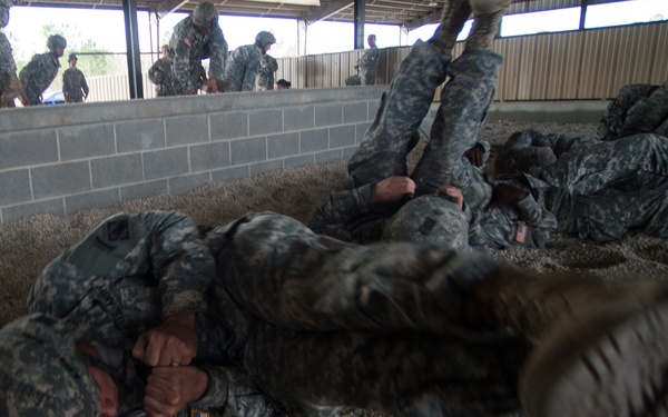 U.S. Army soldiers in ACU (Army Combat Uniform) digital camouflage are engaged in combatives or hand-to-hand combat training inside a covered pit filled with gravel or sand. Multiple pairs of soldiers are grappling on the ground while additional soldiers observe from an elevated position behind a concrete block wall. The image is taken from a low angle, with a boot prominently in the foreground, suggesting dynamic close-quarters action.