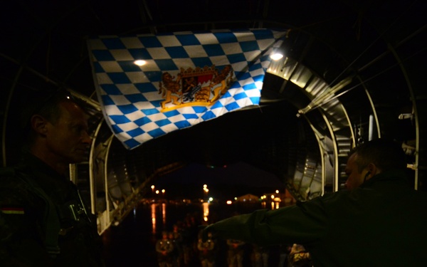 Two military personnel in flight suits are inside what appears to be the rear cargo ramp of a large military aircraft at night. They are holding up a Bavarian flag (blue and white diamond pattern with the Bavarian coat of arms) inside the aircraft. A group of people is visible in the background on the tarmac through the open ramp.