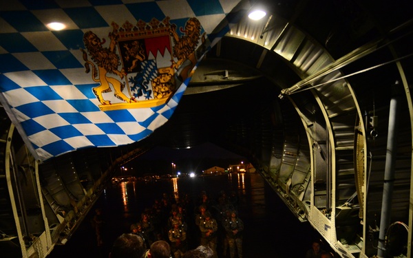 A nighttime scene inside what appears to be a military transport aircraft (likely a C-130 Hercules) with the rear cargo ramp open. Personnel in military camouflage uniforms are gathered inside the aircraft, with the flag of Bavaria (blue and white diamond pattern with coat of arms) prominently displayed at the top of the frame. The scene suggests a military loading or briefing operation at an airfield.