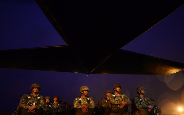 A group of military personnel in combat uniforms and helmets are standing at attention on a wet tarmac at night, positioned beneath the wing of a large military aircraft. The scene appears to be a pre-deployment or staging operation at an airfield, with runway lights reflecting off the rain-soaked surface. The low-light conditions and dramatic composition suggest this is an operational or training deployment scenario.