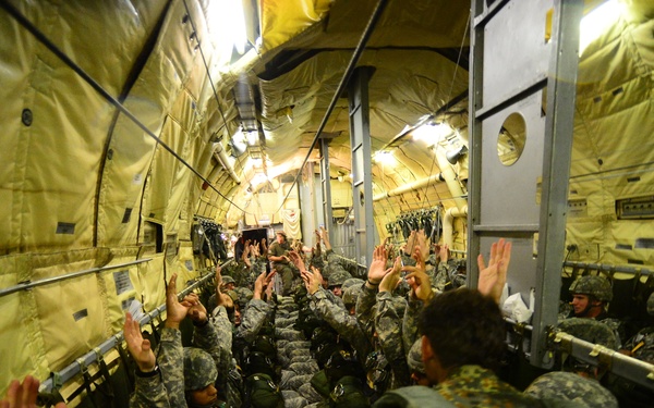 The image shows a large group of military personnel packed inside the cargo hold of a military transport aircraft, likely a C-130 or similar aircraft. The soldiers are wearing camouflage uniforms and helmets, with many raising their hands, possibly during a pre-jump briefing or equipment check for a parachute operation. The interior is illuminated by overhead lighting, revealing the yellow padded walls and structural framework typical of military transport aircraft.