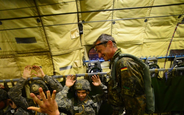 Military personnel are seated inside what appears to be a military transport aircraft, wearing full combat gear and parachute harnesses. A soldier with a visible German flag patch on the right sleeve stands and appears to be instructing or briefing the seated troops. The interior lighting gives a yellow-green cast to the scene, consistent with aircraft interior illumination.