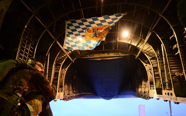 A military personnel in camouflage uniform is crouching near the open rear cargo ramp of a large military transport aircraft, viewed from inside the aircraft looking outward. A blue and white checkered flag, identifiable as the Bavarian state flag, is visible hanging inside the aircraft's cargo hold. The scene appears to be at dusk or dawn on a wet airfield tarmac.