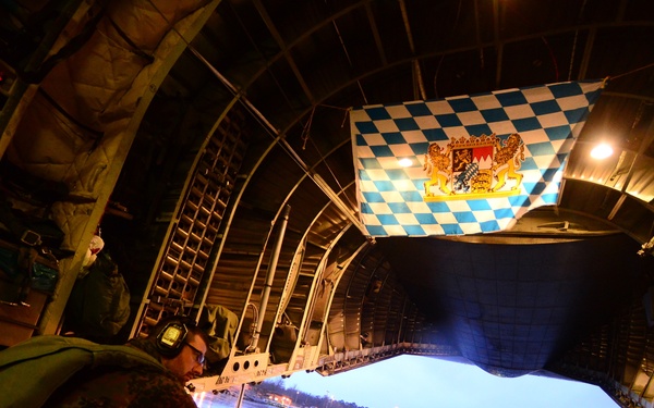 A soldier in camouflage uniform crouches near the open rear ramp of a military cargo aircraft, likely a C-130 Hercules or similar transport, during dusk or dawn conditions. A blue and white checkered flag with a coat of arms — consistent with the flag of Bavaria, Germany — is displayed at the top of the cargo ramp opening. The aircraft appears to be on a wet tarmac at a military airfield with other aircraft and structures visible in the background.