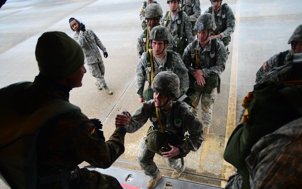 Military personnel in combat uniforms are boarding what appears to be a military transport aircraft via a rear cargo ramp. Soldiers wearing parachute harnesses with yellow static lines are queued on the tarmac, being assisted onto the aircraft ramp by personnel already aboard. The scene suggests a parachute jump operation or airborne training exercise.