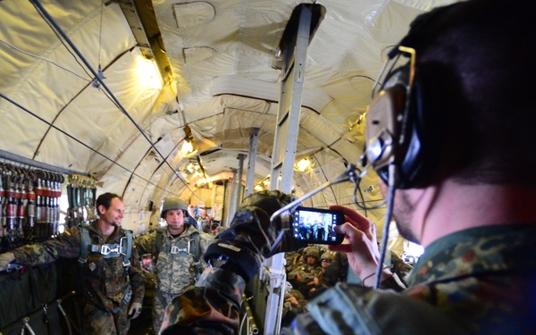 The image shows military personnel inside the cargo hold of a military transport aircraft, equipped with parachute gear, suggesting preparation for a parachute jump operation. Soldiers wearing German Bundeswehr flecktarn camouflage uniforms are visible, with one soldier in the foreground recording the scene on a smartphone. The interior of the aircraft shows static line anchor cables along the fuselage walls, typical of airborne operations.