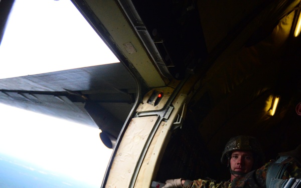 Two military personnel in camouflage uniforms and parachute harnesses are positioned at the open door of a military aircraft in flight. One soldier is leaning toward the door opening, appearing to assess or prepare for a jump, while forested terrain and a body of water are visible far below. The interior of the aircraft is dimly lit with yellow lighting strips visible on the ceiling.