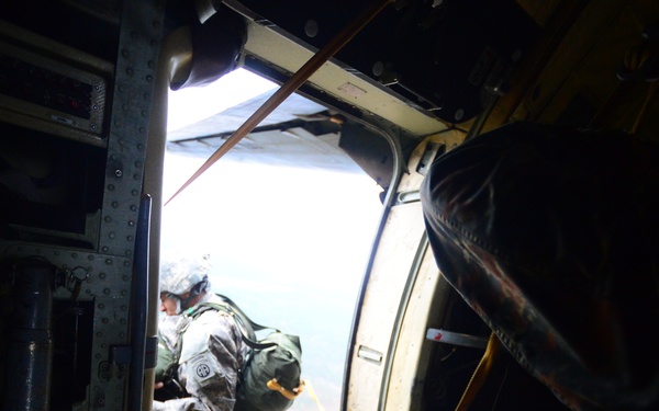 A military paratrooper is visible at the open door of an aircraft, in the process of jumping or preparing to jump. The image is taken from inside the aircraft looking toward the open door, with ground terrain visible far below. The soldier is wearing full combat gear including a parachute pack and appears to have a unit patch on their shoulder.