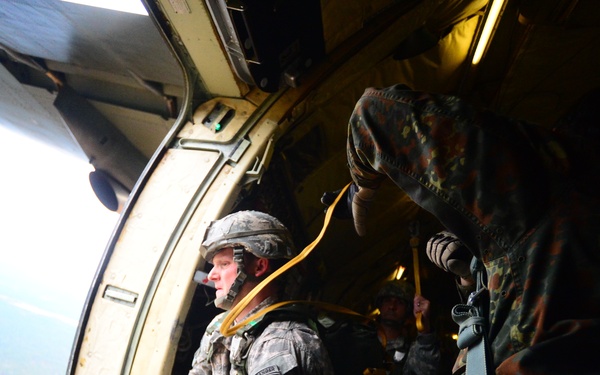 Military paratroopers are positioned at the open door of a military transport aircraft, preparing for a jump. A U.S. Army soldier wearing an 82nd Airborne Division patch is visible near the aircraft door, with a static line attached, alongside soldiers in different camouflage patterns. The ground is visible far below through the open aircraft door.