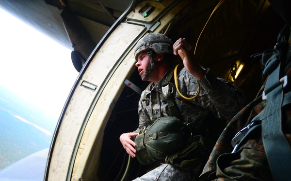 A military paratrooper in US Army ACU (Army Combat Uniform) is positioned at the open door/ramp of a military aircraft, preparing to jump. The soldier is holding a yellow static line and appears to be looking out at the terrain below. A second soldier in what appears to be German Flecktarn camouflage pattern is visible to the right.