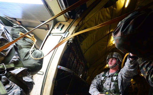 Military paratroopers are conducting an airborne jump operation from what appears to be a military transport aircraft. One soldier is actively exiting the aircraft door while another stands inside observing, with a static line visible connecting jumpers to the aircraft. A third individual in a different camouflage pattern (possibly a foreign military observer) is visible on the right side.