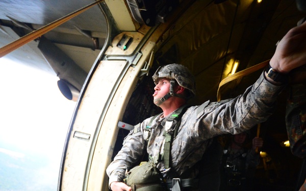 A soldier in US Army ACU (Army Combat Uniform) is positioned at the open door of a military aircraft, appearing to be preparing for or in the process of a parachute jump. The soldier is wearing full jump gear including a helmet, parachute harness, and carrying additional equipment. The terrain visible below shows forested land with a winding river or road.